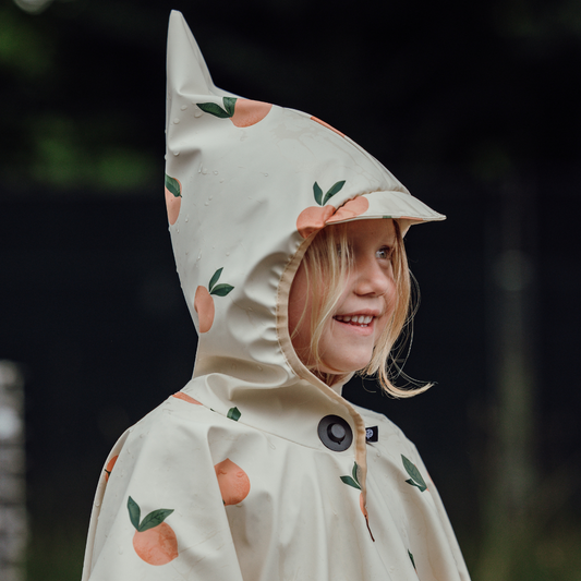 Mädchen trägt wasserfesten Kinderponcho mit Magnetknopf und Orangenprint, Seitenansicht mit Zipfelkapuze.