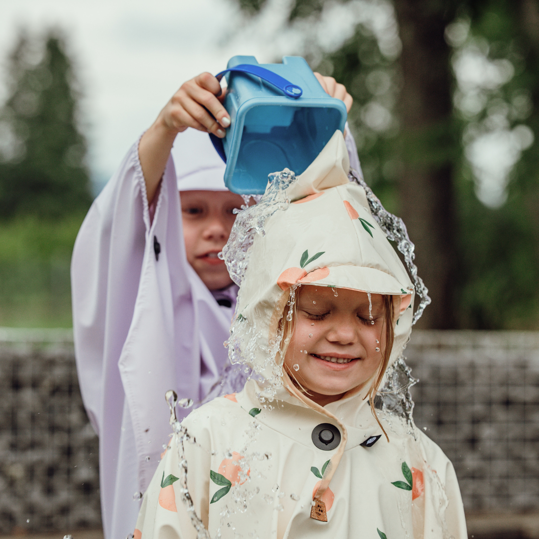 Kind mit Regenponcho und Magnetknopf wird spielerisch mit Wasser überschüttet – hält trocken.