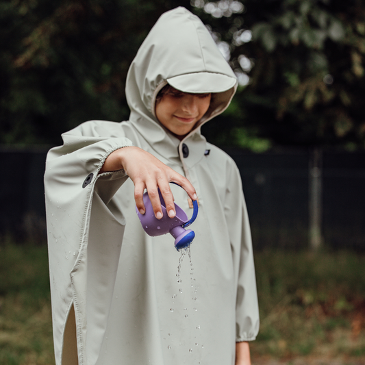 Kind im Regenponcho mit Kapuze und Magnetknopf am Hals – hält Gießkanne und demonstriert wasserabweisendes Material.