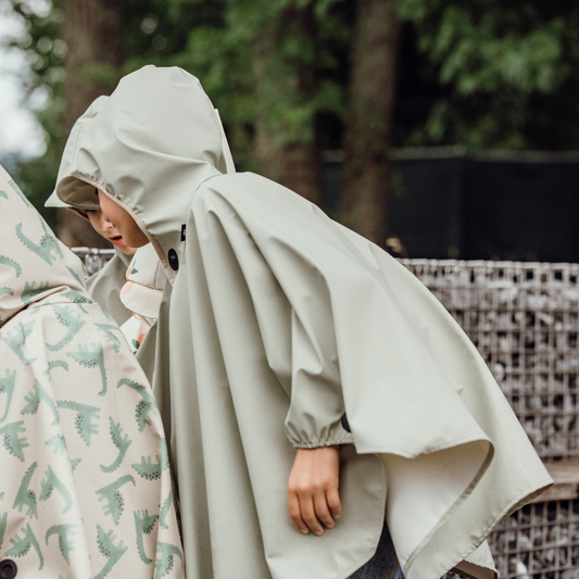 Zwei Kinder im Regenponcho mit Kapuze – Fokus auf großzügigen Schnitt und Magnetknopf an der Seite für einfaches An- und Ausziehen.