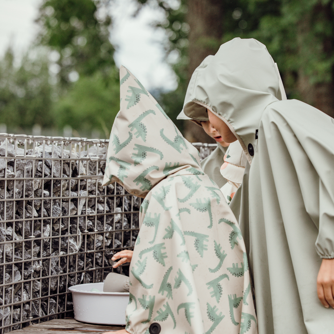 Zwei Kinder im Regenponcho entdecken draußen Wasser – funktionale Outdoorbekleidung mit Magnetknöpfen und Kapuze